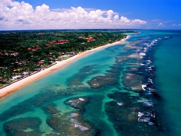 Vista aérea do litoral sul da Bahia, com vegetação, faixa de areia e formações recifais no mar.